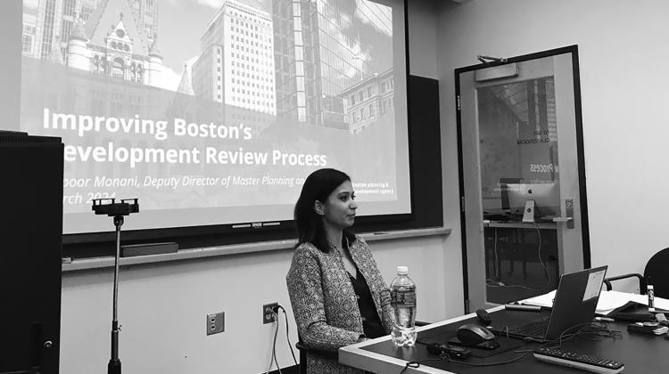 Black and white image of an individual sitting at the head of a table with a slideshow in the background