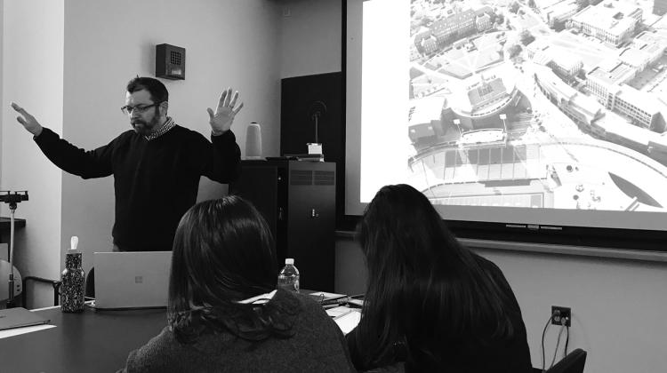 Black and white image of an individual standing in front of a group of students with slide show in the background