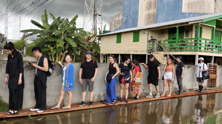 A single file line of individuals standing on a bridge of wood, flanked by water