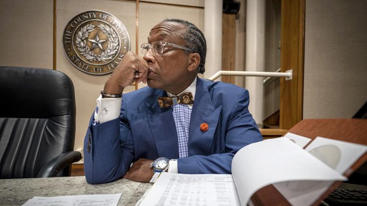 An individual sites at a desk with a spreadsheet open in front of them and the seal of the County of Dallas in the background